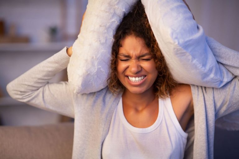 Angry student girl trying to sleep, covering ears with pillows and closing eyes, irritated with noisy neighbors