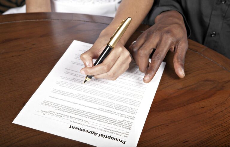 A close-up of two individuals reviewing and signing a prenuptial agreement document with a pen on a wooden table
